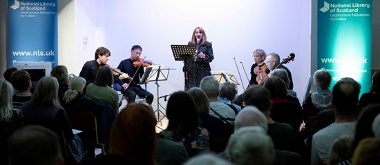 A string quartet spotlighted on stage with one singer in the middle. The backs of heads of the audience is in the foreground.