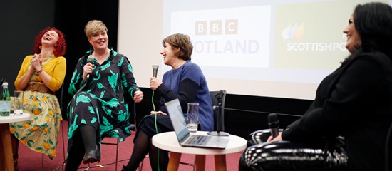 Four women sitting on a stage talking. On the screen in the background is the BBC Scotland logo.